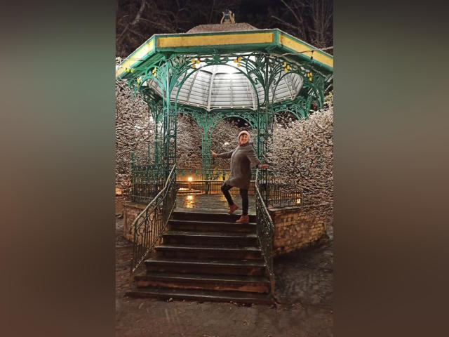 BellisimaAh stands on a green, metal, hexagonal gazebo's steps, wearing a gray coat and red beanie, with her back to the camera, in a snowy, nighttime setting.