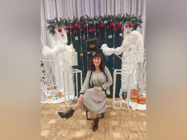 BellisimaAh with red hair, gray dress, and black boots, sitting on a stool, smiling, surrounded by Christmas decorations, white trees, and red ornaments. Boobs visible under dress.