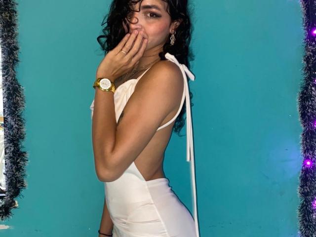 TiffanyFire with curly black hair, wearing a white backless dress, covering her mouth with her hand, wearing a gold watch, standing against a blue wall.