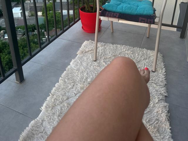 Stefanni's legs, bare with pink toenails, resting on a white shaggy rug, showing her smooth, tan-skinned thighs. A teal cushioned chair with a purple pattern is nearby.