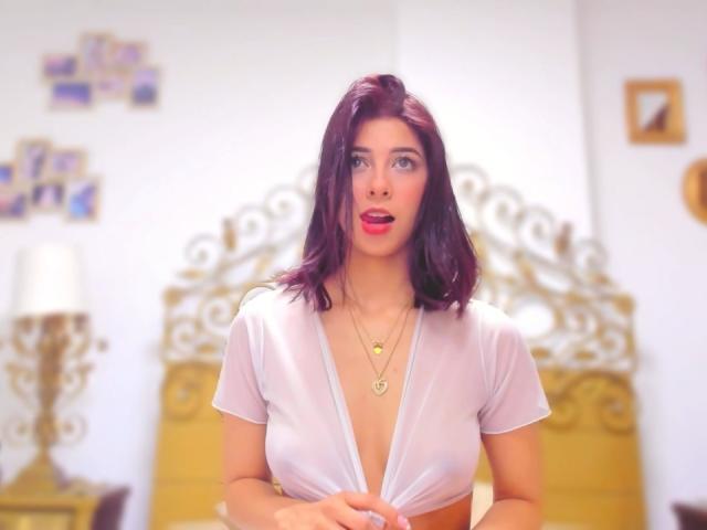 HellenBells with purple hair, white sheer top, exposing her tits, red lipstick, gold necklace, looking up with a slight open-mouth smile, in a brightly lit room.