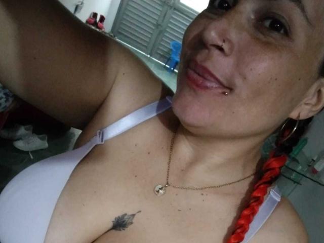 Vennus69 com trança vermelha, sutiã branco, pequeno piercing no nariz, piercing no lábio, pele bronzeada, pequena tatuagem de folha, colar, sorrindo, segurando o celular, mostrando os seios, e uma linha de bronzeamento discreta..