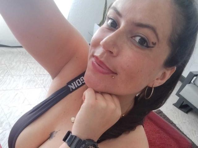 Vennus69, con pechos medianos, cabello negro, aros grandes, piercing en la nariz y piercing en el labio, sonriendo, sosteniendo su barbilla, vistiendo una blusa negra y un reloj negro, en un selfie..