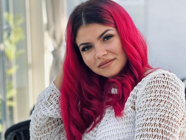 MissSweetty with bright red hair, wearing a white lace top, has a cute smile and dark eyebrows, looking at the camera with a slight head tilt.