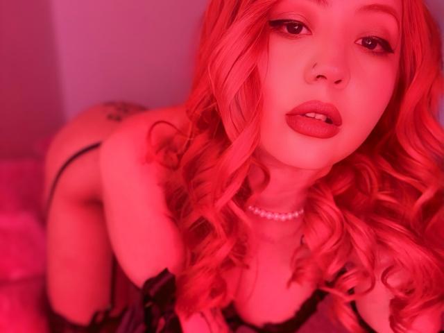 IsabellaLee69 with pink curly hair, red lipstick, black lingerie, pearl necklace, and tattoo on her right butt cheek, kneeling, looking at the camera with a slightly open mouth.