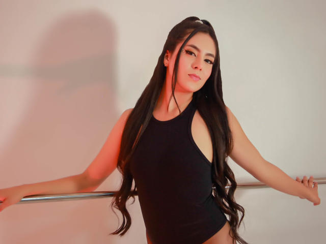 EimmyPark with long black hair, wearing a black sleeveless top, standing with arms on a metal rail, looking down with a slight smile, red light on her right side.