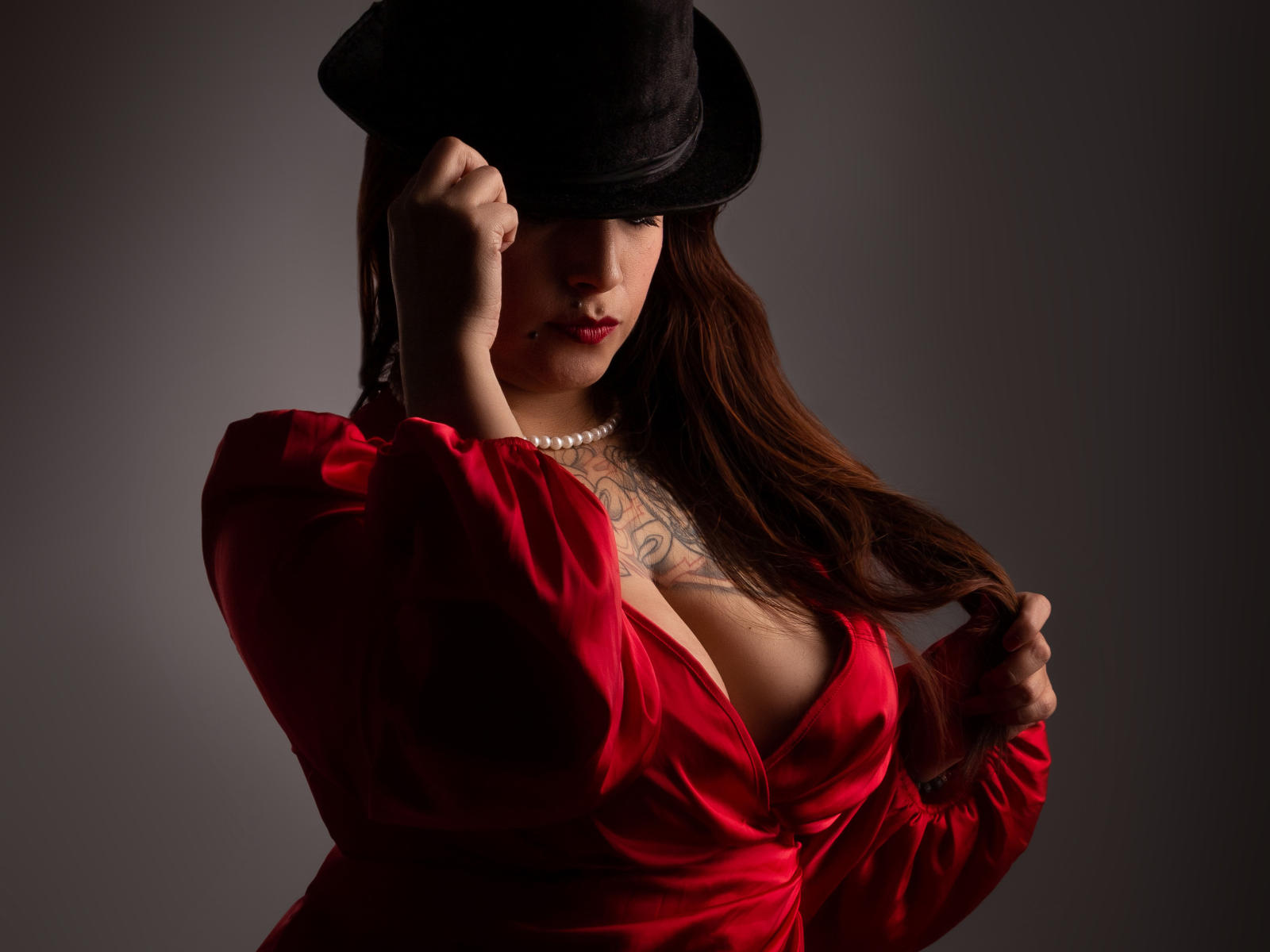 ZoeMichaels with long brown hair, wearing a black top hat, red dress with deep cleavage, pearl necklace, and holding her hat, showing tattoos on her chest.