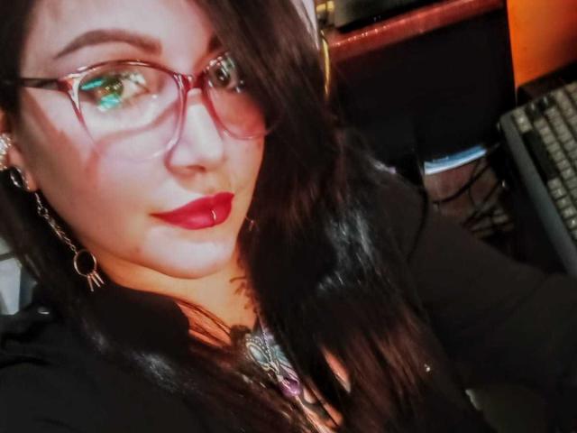 SweetAmeliee con rossetto rosso, occhiali e capelli castano scuro, che mostra il viso e la parte superiore del petto, indossa una maglietta nera e una collana viola, con un leggero sorriso..