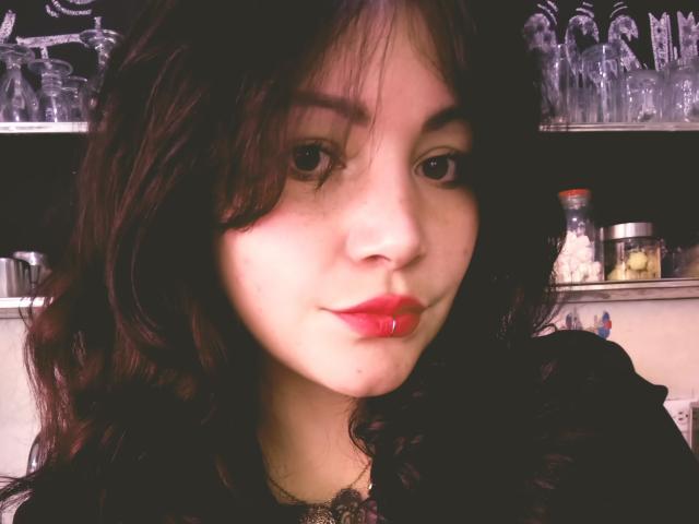 SweetAmeliee, avec ses cheveux bruns foncés ondulés, sa peau claire, son rouge à lèvres rouge et son maquillage discret, regarde l'appareil photo avec un léger sourire, et porte un haut en dentelle noire..