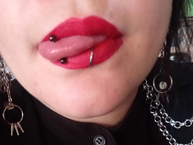 Les lèvres de SweetAmeliee, recouvertes de rouge à lèvres rouge, avec un piercing labial noir et rose, la langue légèrement sortie, portant des boucles d'oreilles noires avec des chaînes, et une chemise noire avec un bouton..