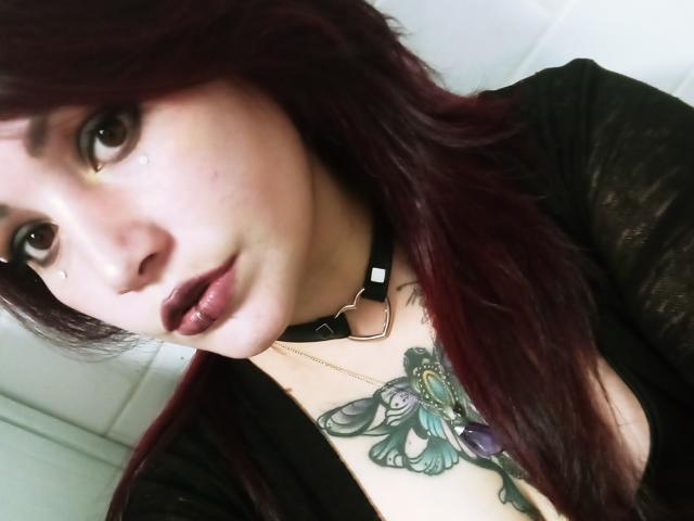 SweetAmeliee, avec ses cheveux roux foncés, son collier noir et son tatouage de papillon sur la poitrine, portant un haut noir, regardant l'appareil photo avec des tatouages en forme de goutte de larme sur les joues, et un rouge à lèvres foncé..