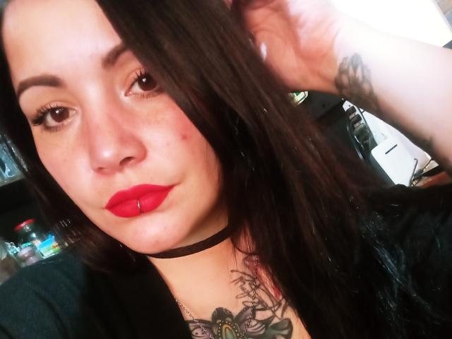 SweetAmeliee, avec de longs cheveux bruns, du rouge à lèvres, un collier noir et un tatouage sur la poitrine, se tient les cheveux, laissant apparaître de petits piercings au nez, et affiche un sourire discret. Ses seins sont petits..