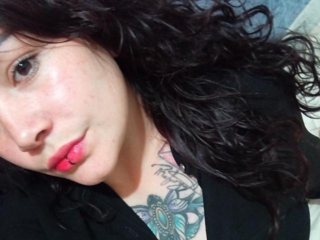 A selfie of SweetAmeliee with curly black hair, pale skin, pink lips, and a colorful tattoo on her chest showing a blue and green floral design.
