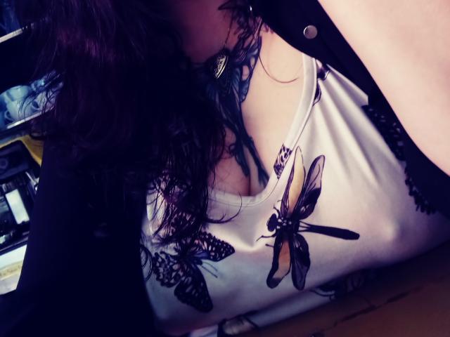 La canotta bianca di SweetAmeliee con stampe di farfalle e merletti, che mostra i suoi seni, un reggiseno nero in pizzo e i suoi capelli scuri e ricci, parzialmente coperti da una camicia nera..