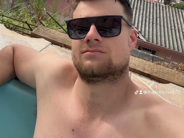 Tesouro, sin camisa y con barba, lleva gafas de sol negras, tiene un pecho musculoso y una expresión relajada. Su brazo está apoyado sobre una superficie azul..