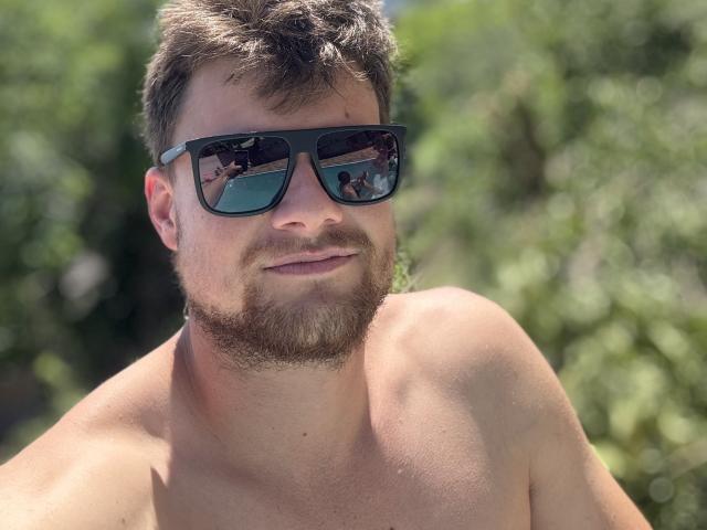 Tesouro with a beard, wearing black sunglasses, shirtless, showing a slight smile, with a reflection of a building in the sunglasses, and a blurred green background.