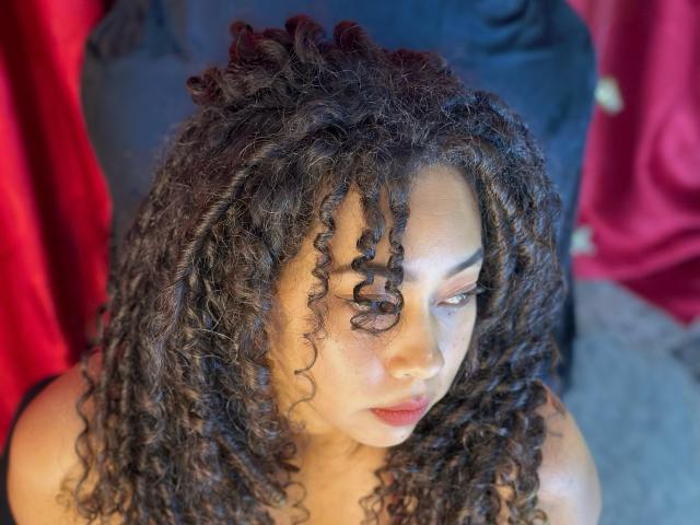 RebeccaMils with curly brown hair, looking to the side, has a pensive expression, red lipstick, and a nose piercing, wearing a black top. Red and blue background.