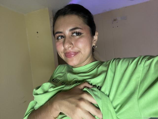 MaiaHairy with dark hair, green shirt, and gold earrings, holding her shirt, smiling, pink lips, small nose, and dark eyeliner, in a dimly lit room.