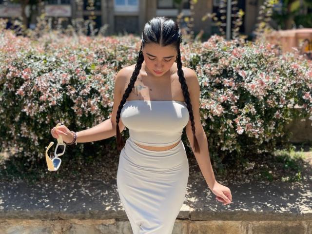 SarahWrigh with long black braids, white strapless top, and white skirt, holding sunglasses, showing her tits and ass, standing in front of a bush with pink flowers.