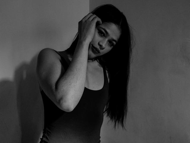 EuphoriaRush, com cabelo preto e liso, vestindo uma regata preta, tocando a cabeça com uma expressão levemente irritada e sedutora, em uma foto em tons de cinza, cheia de sombras..