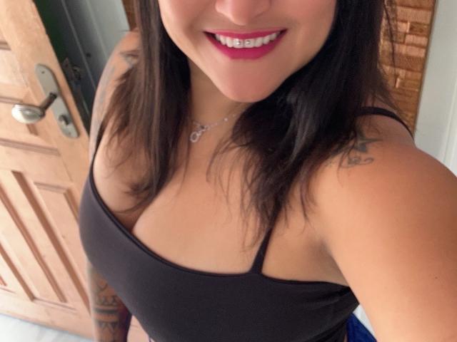 StarHot69 with pink lipstick, smiling, brown tank top, visible tattoos on shoulder, necklace, brown hair, medium tits, brown door in background.