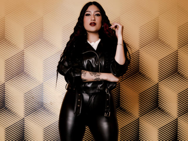 EmmaRosseti with long black wavy hair, dark red lipstick, black leather pants, and jacket, showing tattooed arm, standing, looking at camera, hand near face.