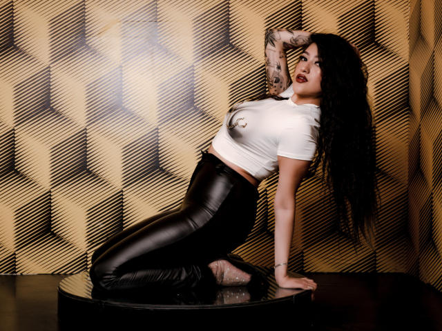 EmmaRosseti with long black curly hair, white tee, and black leather pants, kneeling on a black platform, arms raised, showing tattoos, and looking seductively at the camera.