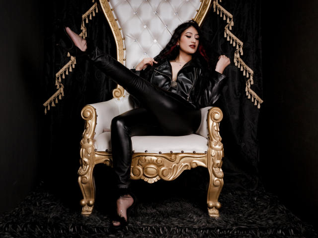 EmmaRosseti with black leather jacket, black pants, and pink heels, legs spread on an ornate gold and white chair, dark red lipstick, long black hair, and confident expression.