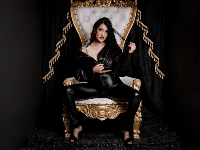 EmmaRosseti sits on a gold and white throne, wearing black leather pants and a zipped, partially open black leather jacket, showing her tits. Her black hair is loose.