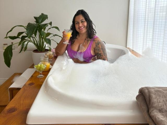 StarHot69 with big tits in a white bathtub, covered in bubbles, holding a glass of orange drink, smiling, black curly hair, tattooed arm, purple bra, wooden table.