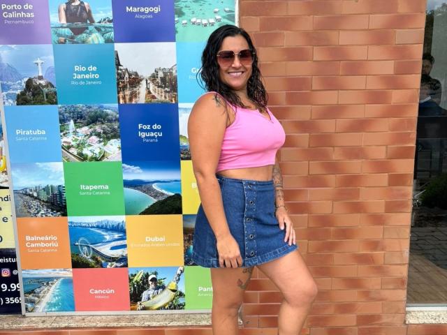 StarHot69 has tan skin, dark wet hair, sunglasses, pink crop top, and denim skirt, standing against a brick wall with a tattoo on her left thigh. Boobs visible.