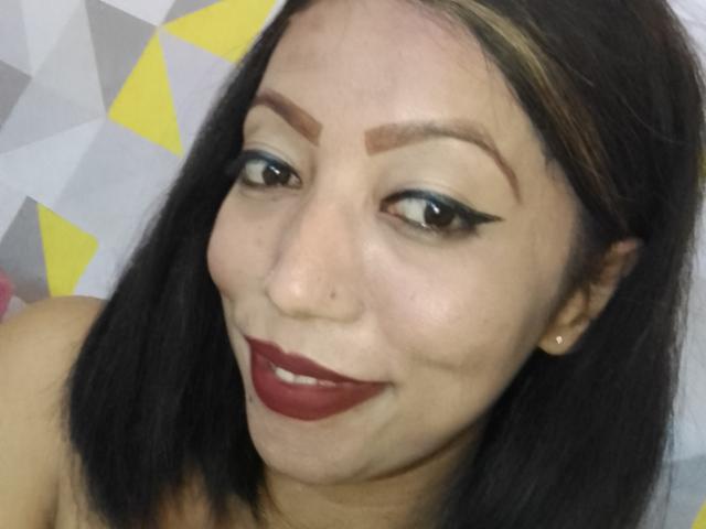 Anniatattoo has dark brown hair, thick eyebrows, black eyeliner, red lipstick, and a nose ring, smiling with a cute face and small tits in the photo.