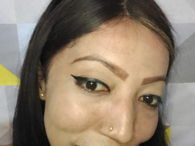 AnniaTattoo has brown eyes, black eyeliner, thick eyebrows, a nose piercing, and long brown hair. She's smiling with a slight pout. Her skin is light with a subtle tan.