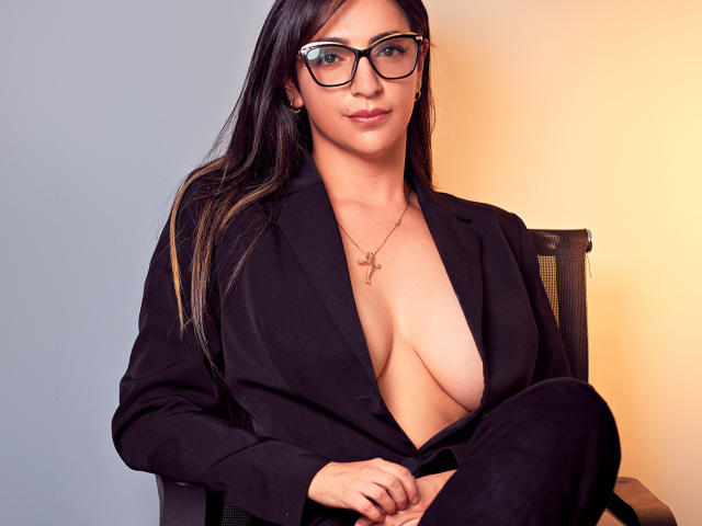 CarolinaaGrey with glasses, black blazer, and deep cleavage, showing her big tits, sitting, looking at camera, long brown hair, cross necklace, slight smile, warm light.