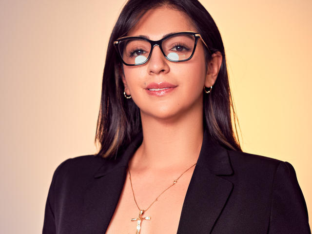 CarolinaaGrey with dark straight hair, black glasses, and a black blazer, wearing gold earrings and a cross necklace, smiling slightly. She's got shiny lips and a cute face.