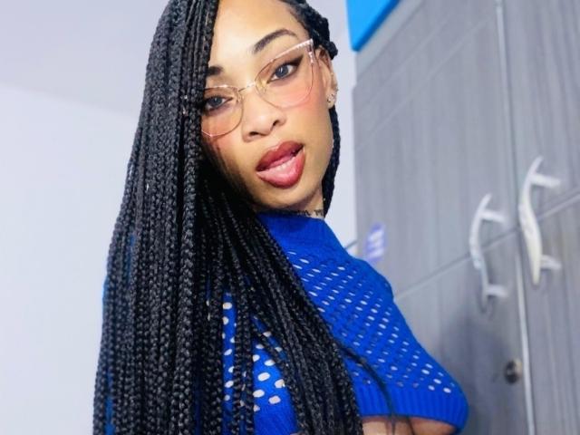 ElaHott, avec de longues tresses noires, portant des lunettes roses et un haut en mesh bleu, regardant en arrière avec des lèvres rouges, dégageant une aura confiante, sexy et élégante..