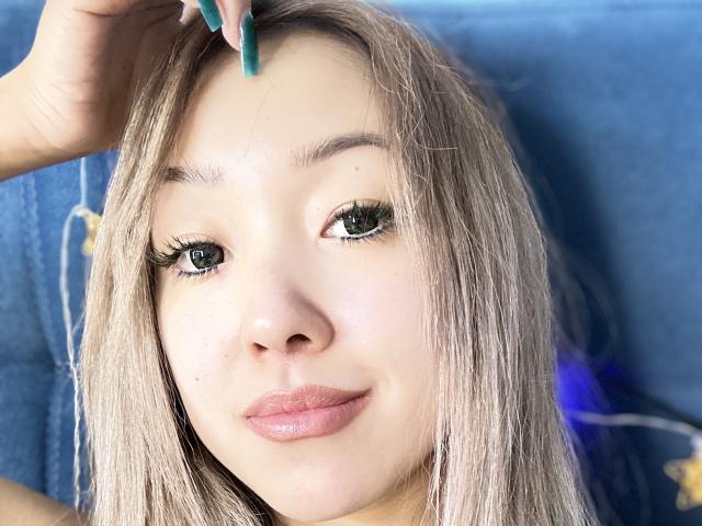 RenyLime has light blonde hair, green eyes with long lashes, and pink lips. She's holding a teal pencil near her forehead, looking at the camera with a slight smile.