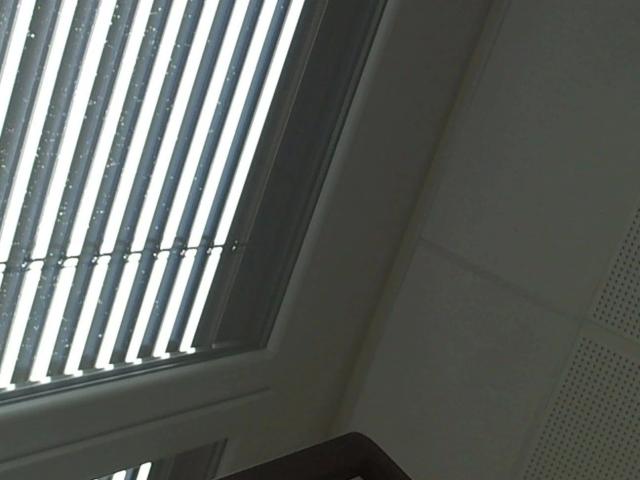 A window with white blinds, letting in bright light, is partially open. The window frame is white, and there's a dark surface below. FlipFlops-hot is not visible.
