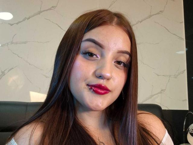 MaylinSanchez has brown hair, red lipstick, and a slightly chubby face with a small nose, looking at the camera with a slight smile. She's wearing a white off-shoulder top.