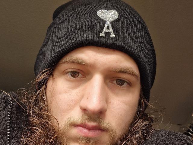 ScxRease hat ein schmutziges Gesicht, braune Augen und einen kleinen Schnurrbart, trägt eine schwarze Mütze mit einem glitzernden Herz und einem "A", langes, braunes Haar und eine dunkle Jacke..