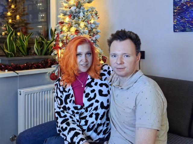 GenrikhBecca with bright orange hair, leopard print jacket, and red shirt, sitting next to a guy with short brown hair, gray polo, and blue jeans, in front of a Christmas tree.