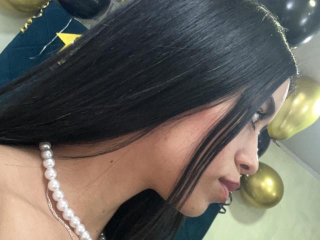 LaurentRay's side profile with long black hair, wearing a pearl necklace, showing a nose piercing and a slight smile. Boobs not visible. Background includes gold balloons.