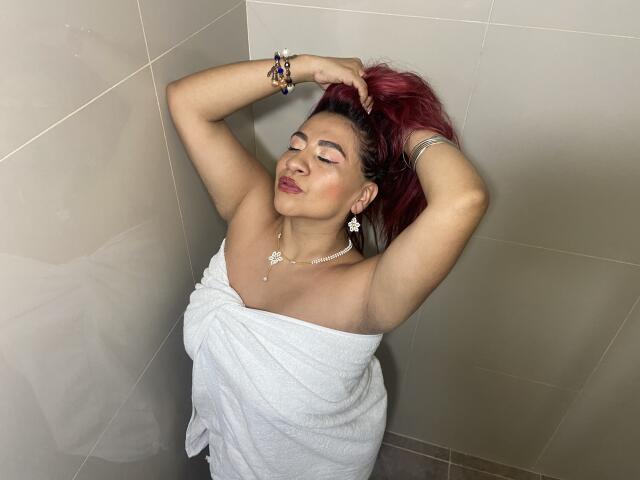 NahelaBigMelonsX with red hair, eyes closed, arms up, white towel covering her tits and ass, standing in a tiled shower, wearing jewelry, looking relaxed and content.