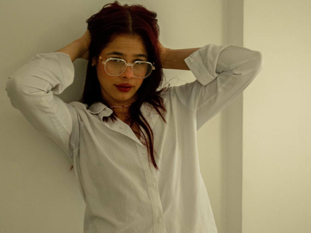 EuphoriaRush met donkerrood haar, een bril, een wit shirt, handen in haar, licht glimlachend, zacht licht, bleke huid, kijkt naar de camera, draagt rode lippenstift, ontspannen pose..