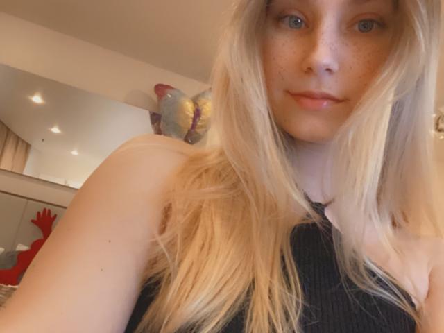 HotCaramells with long blonde hair, freckles, and blue eyes, wearing a black sleeveless top, has a slight smile, and a butterfly tattoo on her upper back.