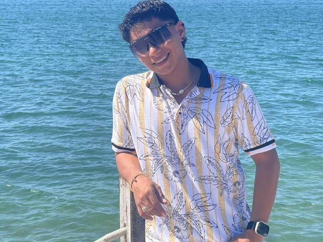 RichiMontes, vestindo uma camisa branca e amarela com estampa de folhas, uma gola preta, óculos de sol e um relógio, está à beira do mar azul, sorrindo e com as mãos nos bolsos..