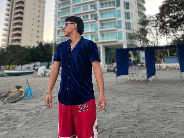 RichiMontes, gekleed in een blauw, fluweelachtig shirt en rode shorts met een wit en rood logo, met zwart haar en een bril, staat op een zandstrand. Op de achtergrond staan fietsen en blauwe zeildoeken..