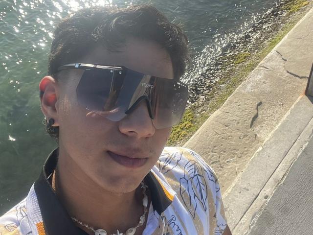 RichiMontes, portant de grandes lunettes de soleil réfléchissantes, une chemise blanche avec des motifs jaunes et un collier de perles, se tient au bord de l'eau, avec de l'eau illuminée par le soleil et une surface en pierre..