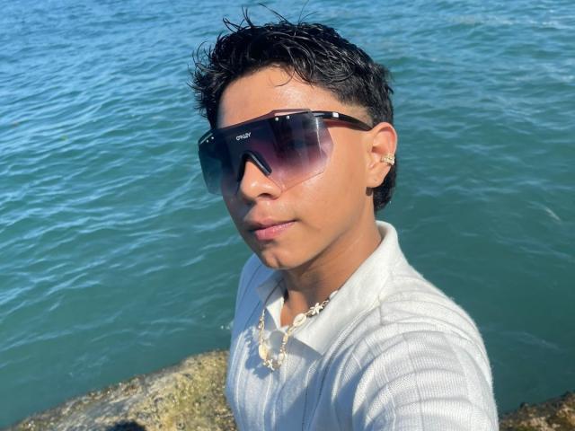 RichiMontes tiene el cabello oscuro y húmedo, lleva gafas de sol moradas grandes, una camiseta blanca con textura vertical, y un collar de concha marina, y está de pie junto a un agua azul con una costa rocosa..