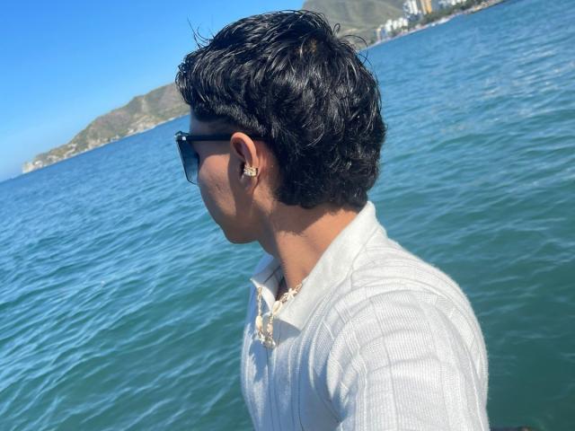 RichiMontes, con cabello negro corto, usando gafas de sol negras, una camiseta blanca con textura vertical y un collar dorado, está de pie junto al océano azul. Tiene un pequeño aro dorado en la oreja..